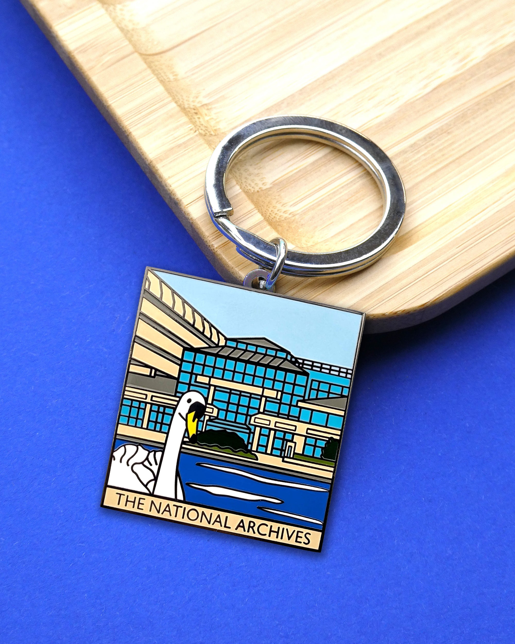 A square metal keychain featuring an illustrated building and swan, attached to a silver split ring, photographed on a blue surface with a light wood board in the background. 